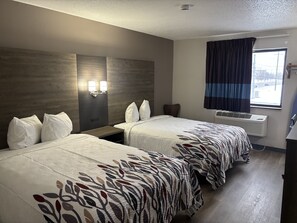 A hotel room with two beds, a nightstand, a wall-mounted lamp, a window with curtains, and an air conditioning unit. - Red Roof Inn & Suites Hammond, IN (Hammond)