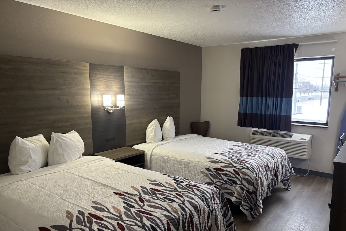 Une chambre d’hôtel avec deux lits, une table de chevet, une lampe fixée au mur, une fenêtre avec des rideaux et un appareil de climatisation.