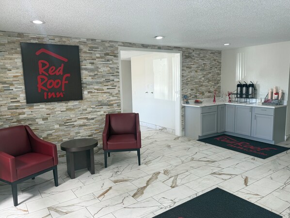 Une chambre d’un Red Roof Inn avec un mur en pierre, deux chaises rouges, une petite table, un coin cuisine avec des placards et une porte donnant sur une autre pièce.