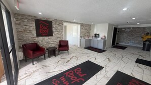 Ein Red Roof Inn Loungebereich mit einer Steinwand, zwei roten Sesseln, einem kleinen Tisch, einer Küchenzeile und einer Person in einem gelben Hemd.