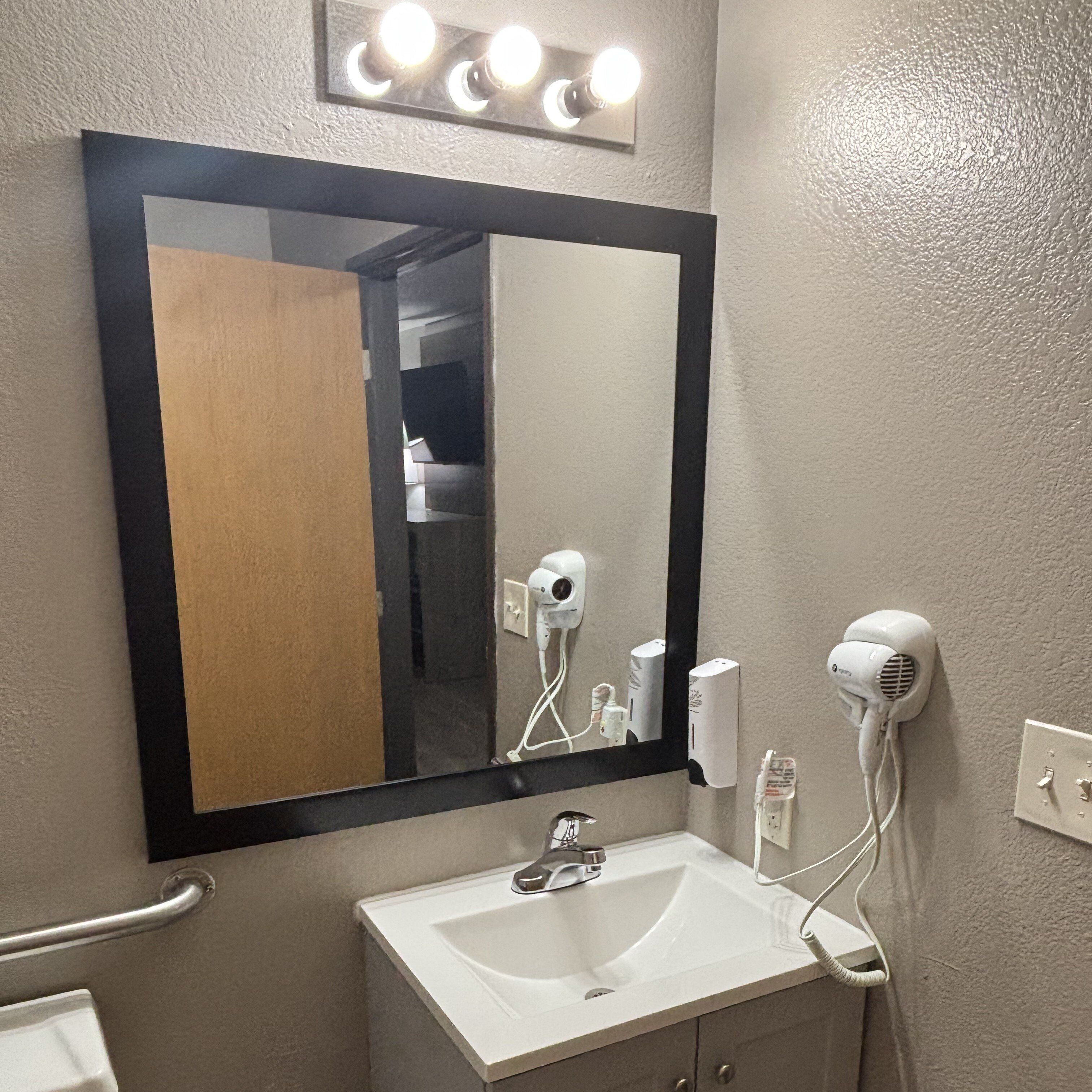 Une salle de bain avec un lavabo blanc, un miroir avec éclairage intégré et deux sèche-cheveux fixés au mur.