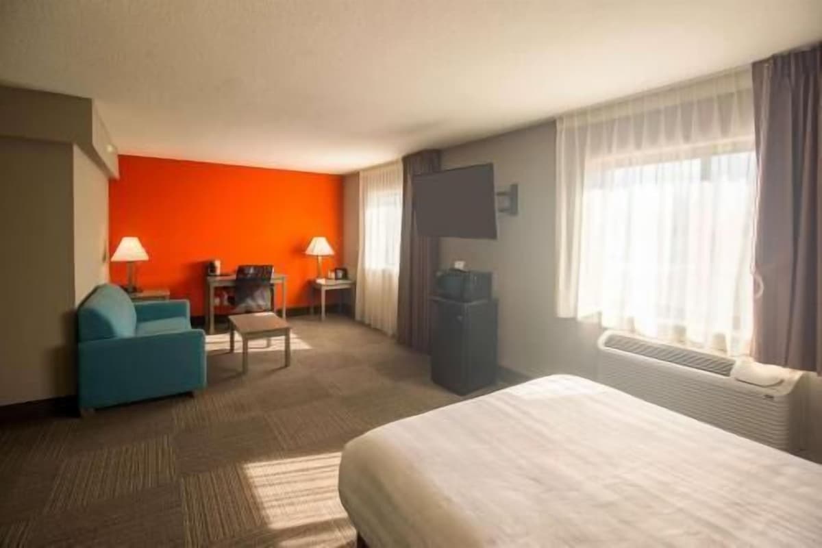 Una habitación de hotel con cama, escritorio, silla, lámpara y ventana con cortinas.