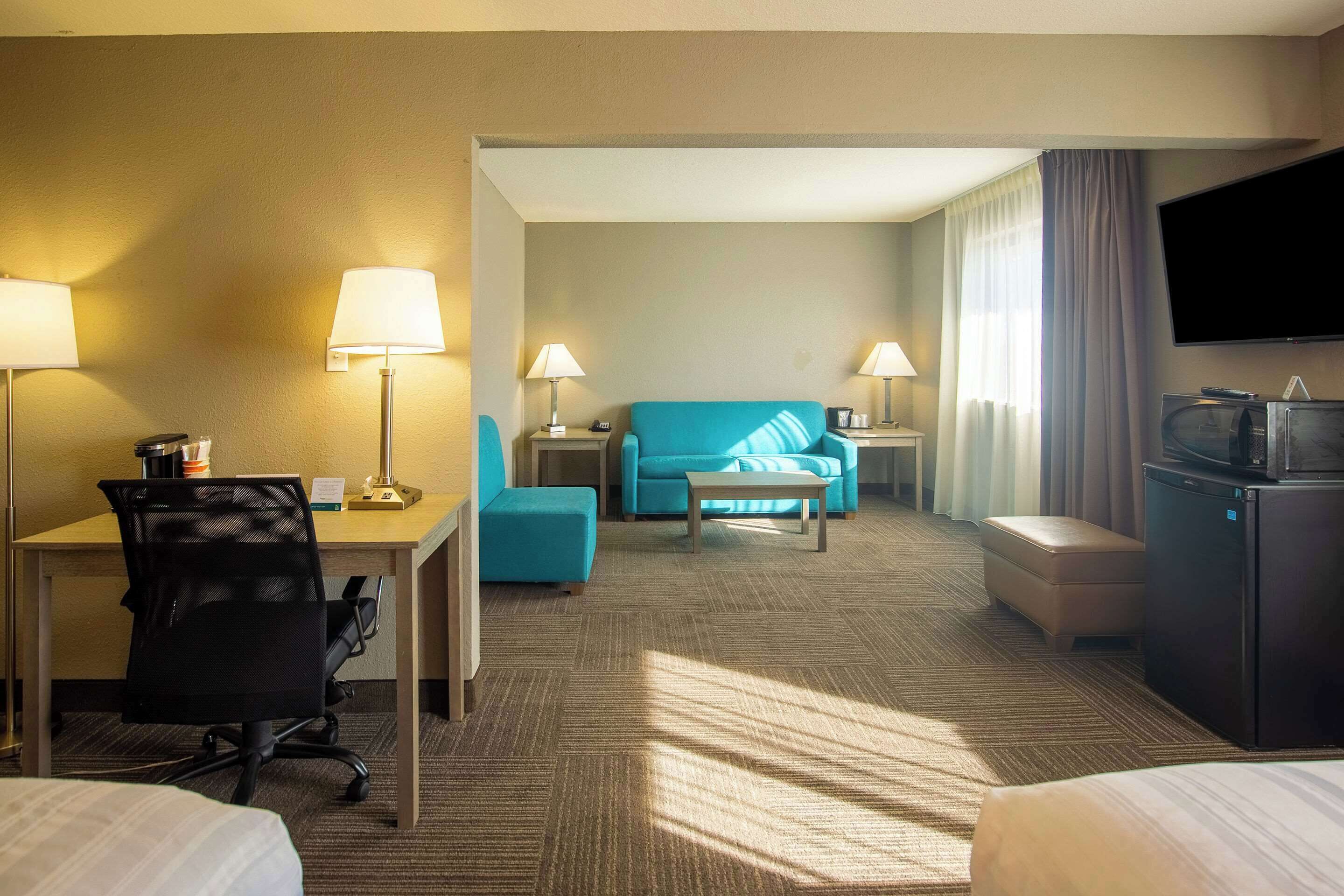 Une chambre d’hôtel équipée d’un bureau, de deux lampes, d’un canapé bleu, d’une petite table, d’une télévision et d’un réfrigérateur.