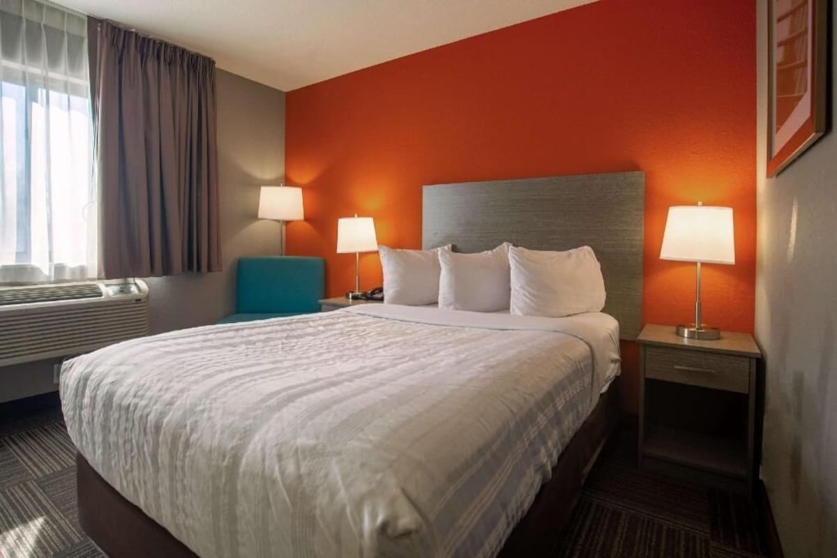 Una habitación de hotel con una cama, dos mesitas de noche, un ventanal con cortinas y un calefactor en la pared.