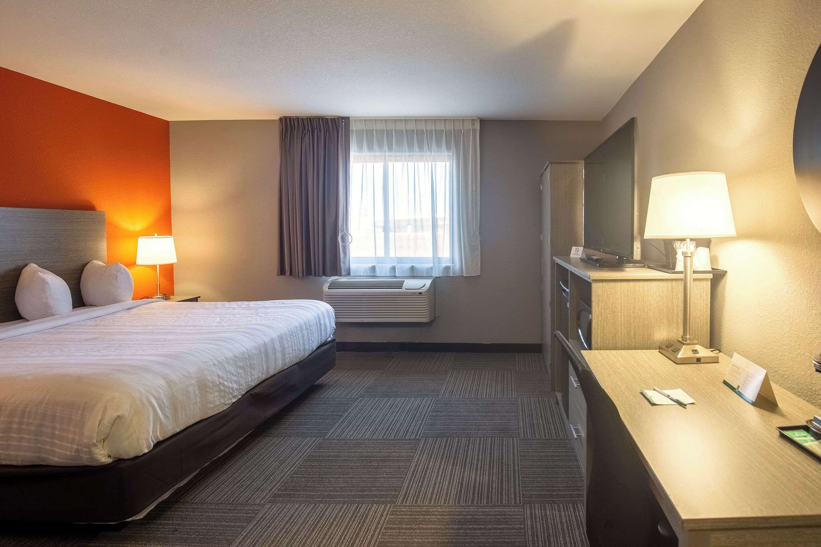 A hotel room with a bed, bedside lamps, a desk with a lamp, a flat-screen TV, and an air conditioning unit.