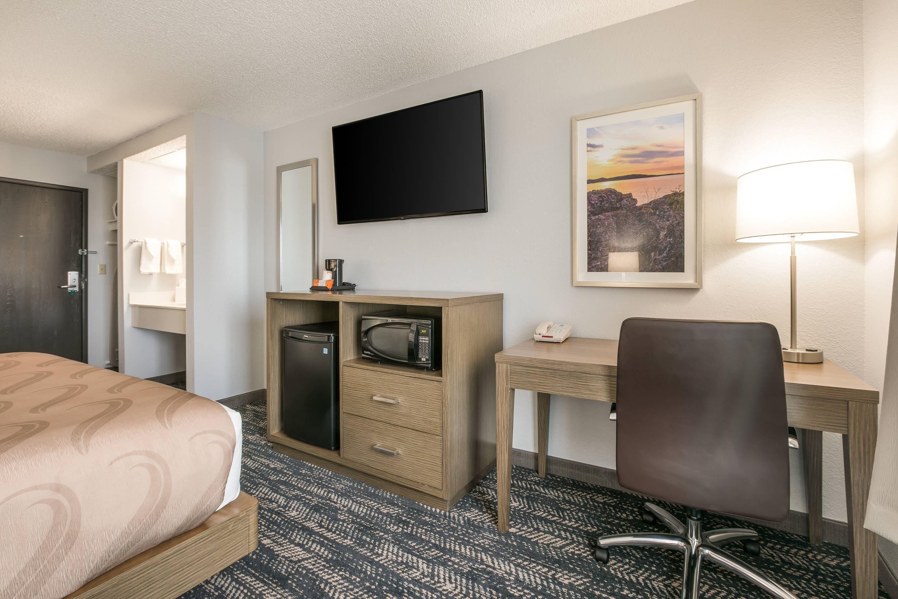 A hotel room with a bed, a desk with a chair, a flat-screen TV, a microwave, a telephone, and a framed picture on the wall.