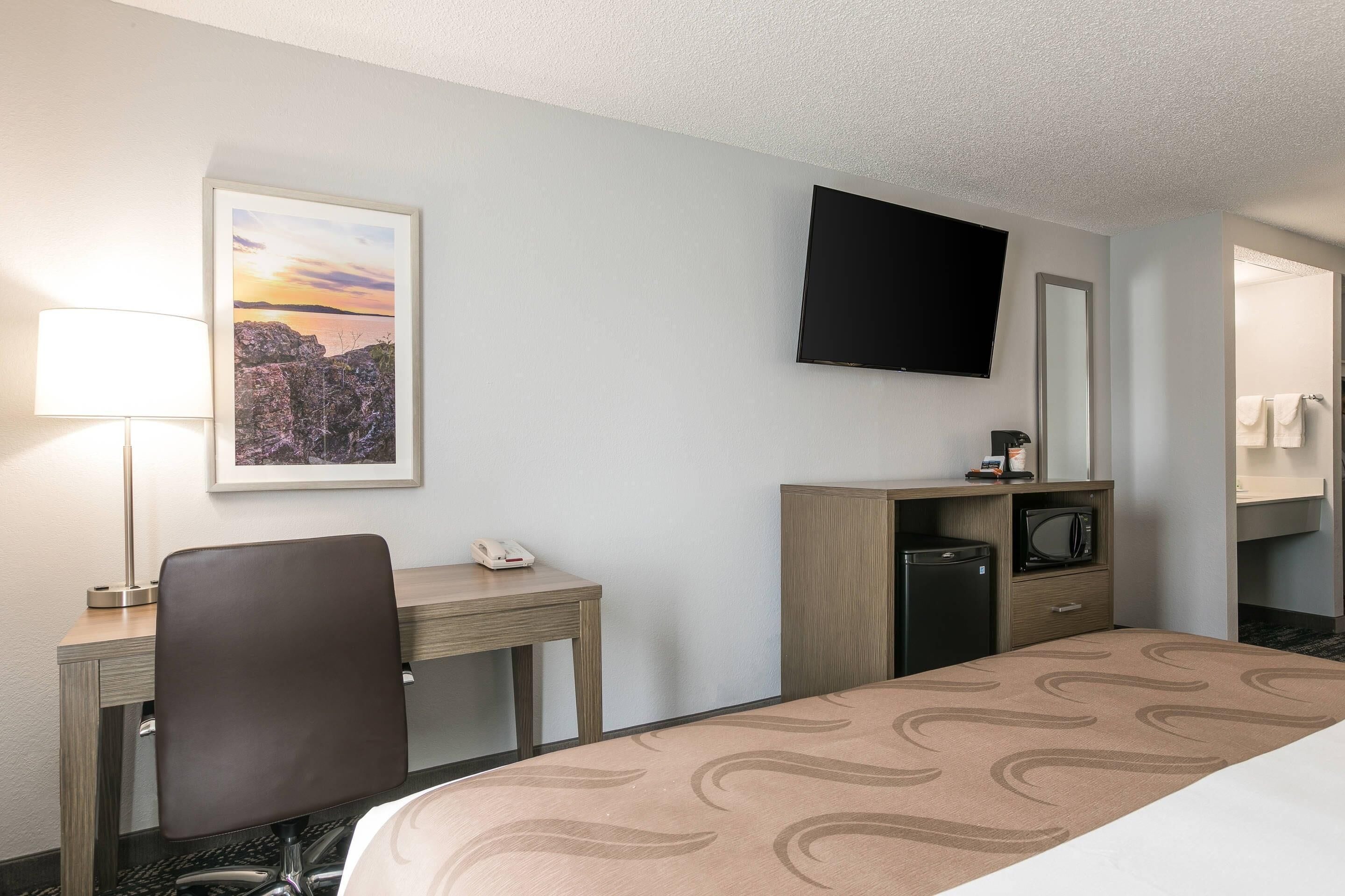 A hotel room with a bed, desk, chair, TV, and a small kitchenette.