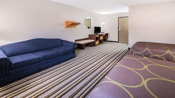 Ein Hotelzimmer mit blauem Sofa, einem Bett mit gemustertem Überwurf, einem Schreibtisch mit Computer, einem Spiegel und einem Streifenmuster-Teppich.