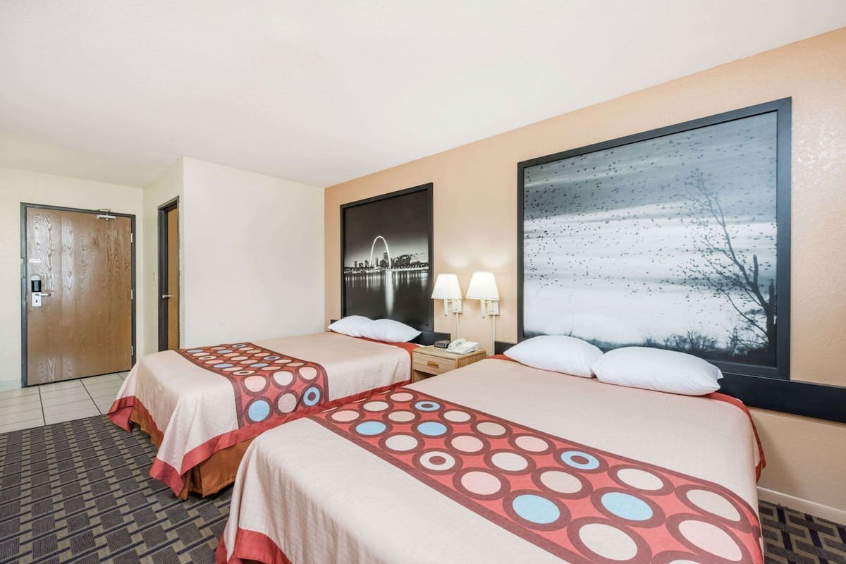 A hotel room with two beds, a nightstand, a cityscape painting, and a window with a snowy landscape view.