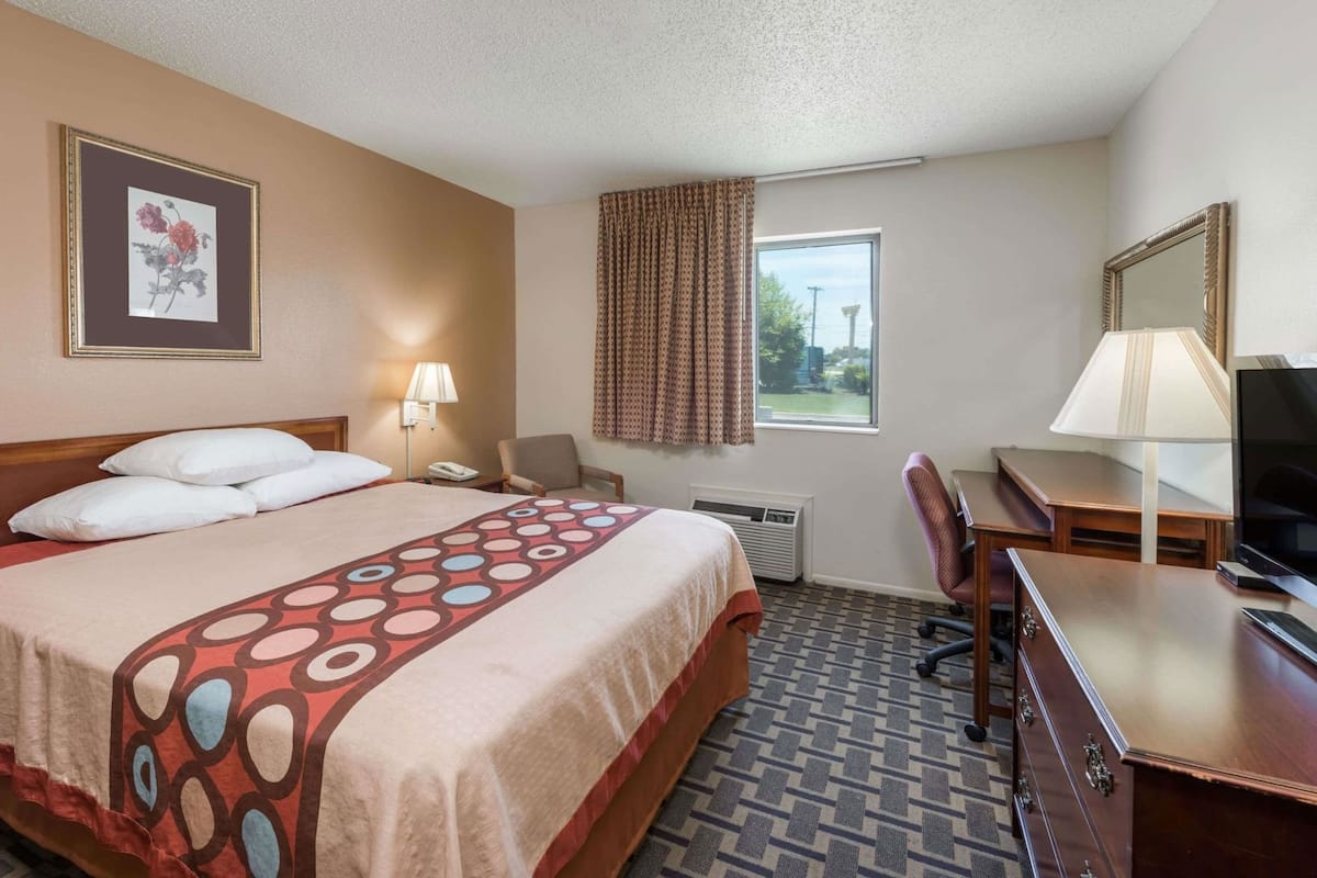 A hotel room with a bed, a desk, a chair, a TV, a lamp, a phone, a window with curtains, and a framed picture on the wall.