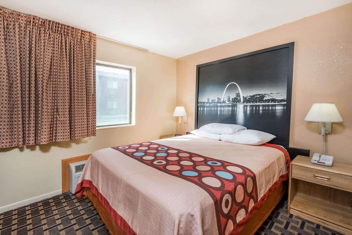 A hotel room with a bed, a window with curtains, a wall-mounted lamp, and a cityscape poster.