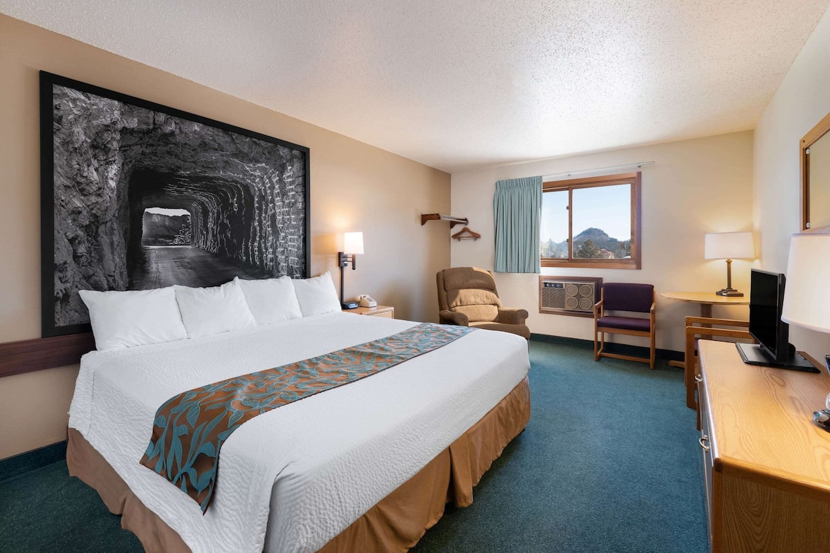 A hotel room with a large bed, a desk with a computer, a chair, a TV, a window with a view, and a large abstract painting on the wall.
