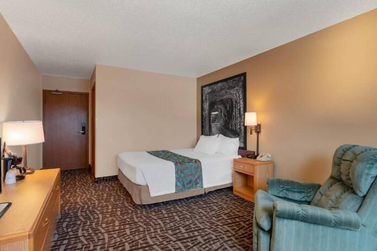 A hotel room with a bed, a desk with a computer, a chair, a wall-mounted TV, and a painting.
