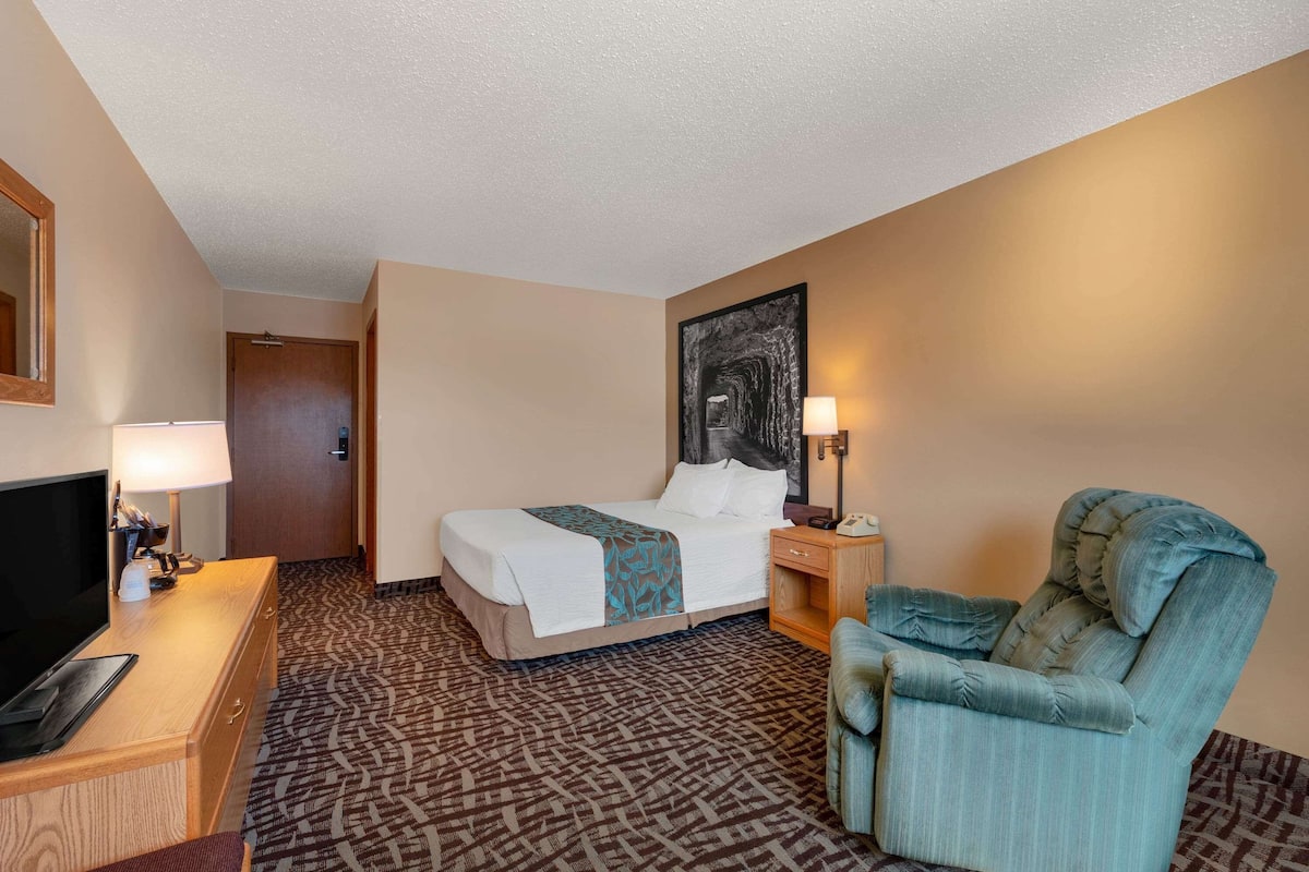 A hotel room with a bed, a desk with a lamp, a chair, and a painting on the wall.