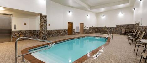 An indoor swimming pool with a stone wall, a wooden deck, and a seating area with chairs.
