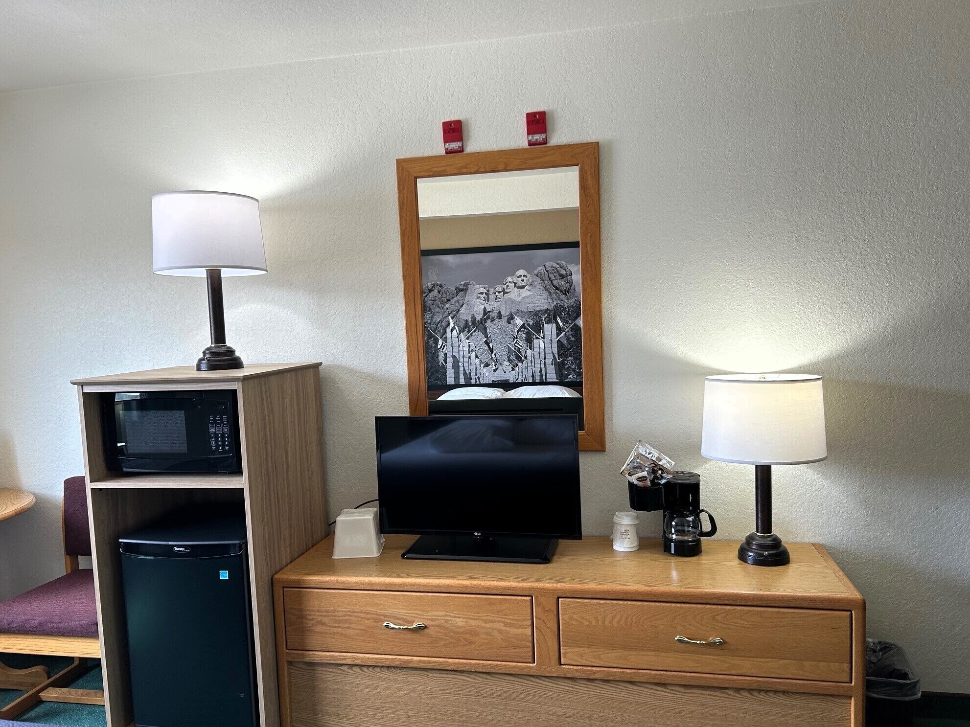 A hotel room with a wooden desk, a television, a microwave, a coffee maker, and a framed picture on the wall.