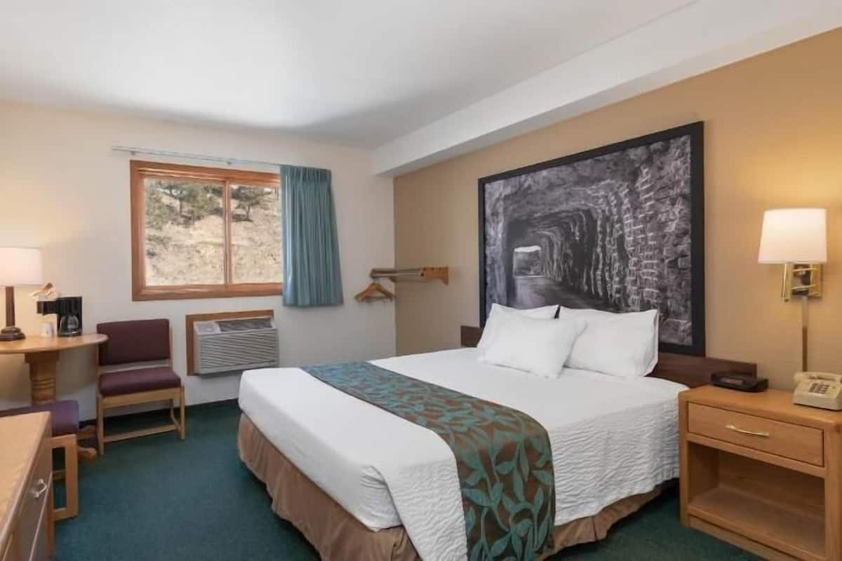 A hotel room with a bed, a desk with a TV, a chair, a window with curtains, a large painting, and a bedside table with a lamp.