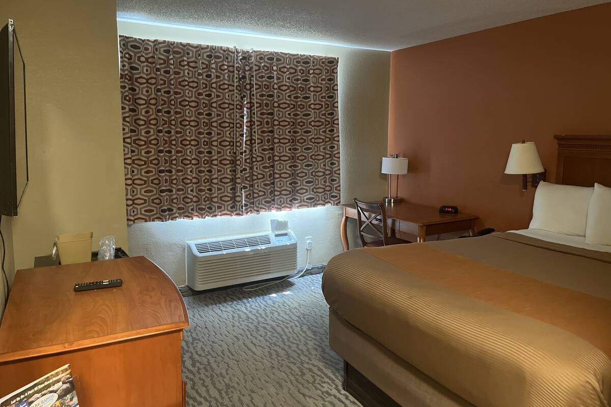 A hotel room with a bed, a desk, a chair, a lamp, a clock, and a window with patterned curtains.