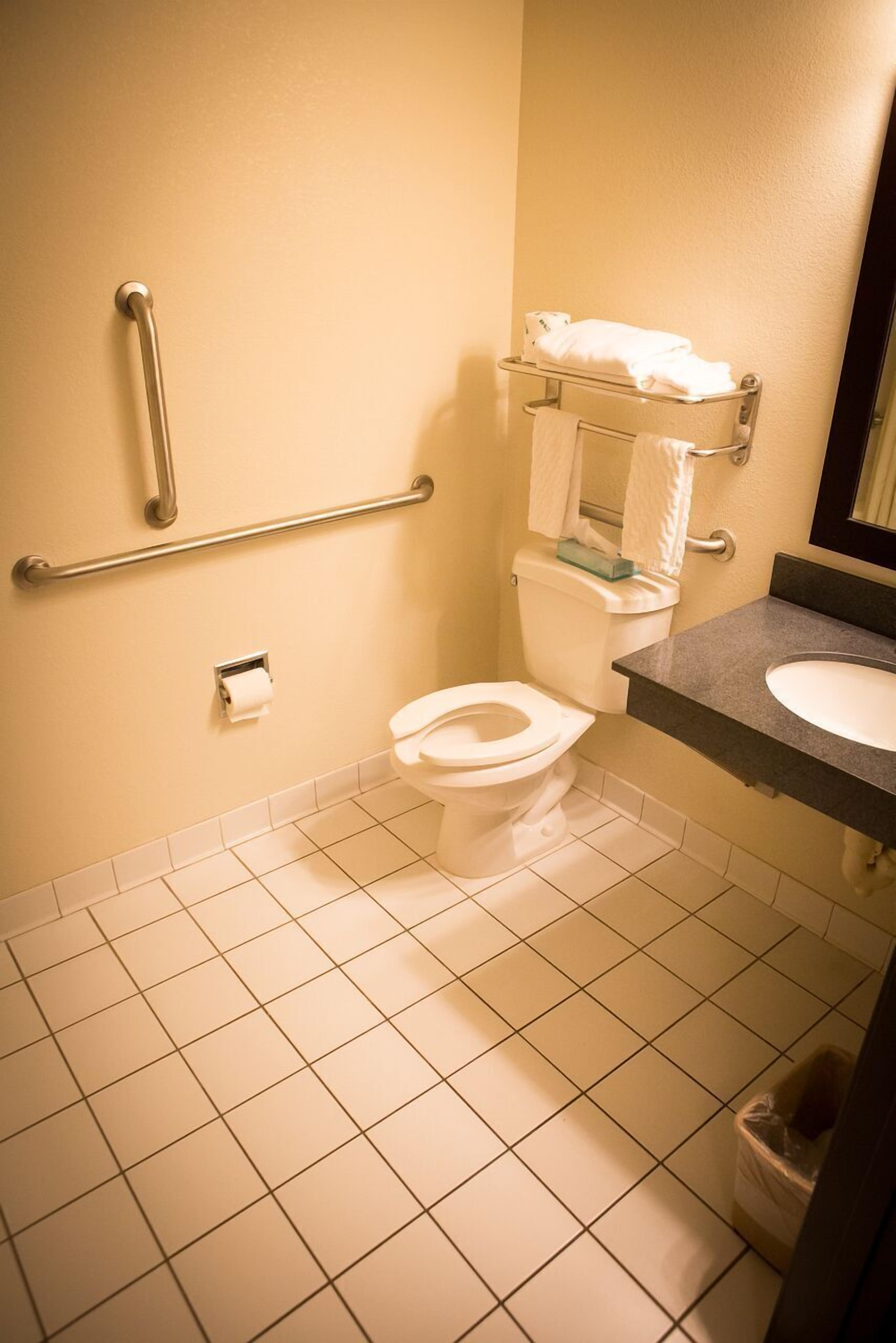 A standard hotel bathroom with a toilet, grab bars, a sink, and a trash bin.