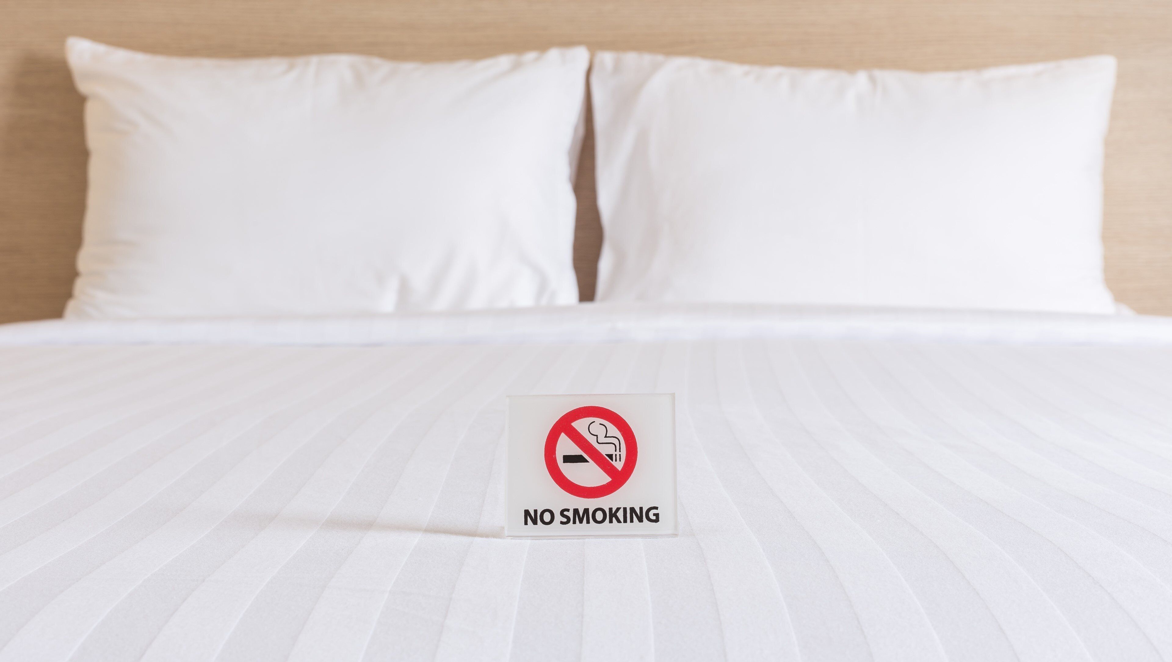 A bed with a white sheet and two white pillows. In front of the bed, there is a sign that reads "NO SMOKING".