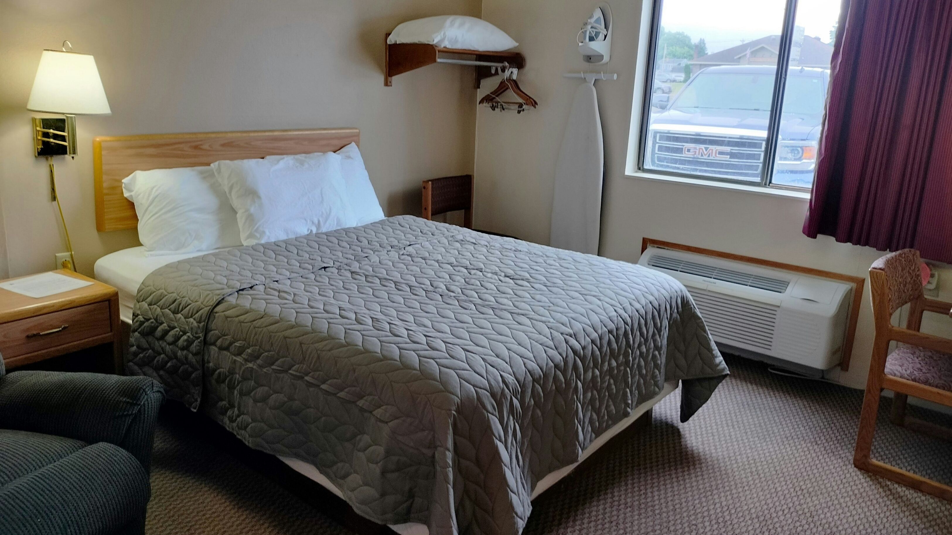 A hotel room with a bed, a chair, a lamp, a heater, and a window with a view of a vehicle.