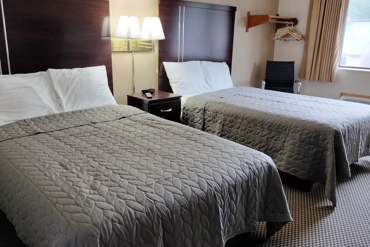 Une chambre d’hôtel avec deux lits, chacun recouvert d’une courtepointe grise matelassée, une table de chevet avec une lampe et un radiateur fixé au mur.