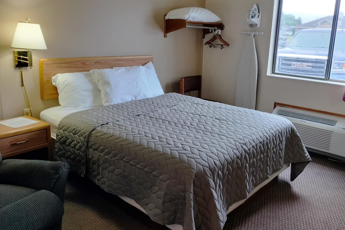 Une chambre d’hôtel comprenant un lit, une table de chevet, une lampe, une chaise, une fenêtre donnant sur un parking et un appareil de climatisation.