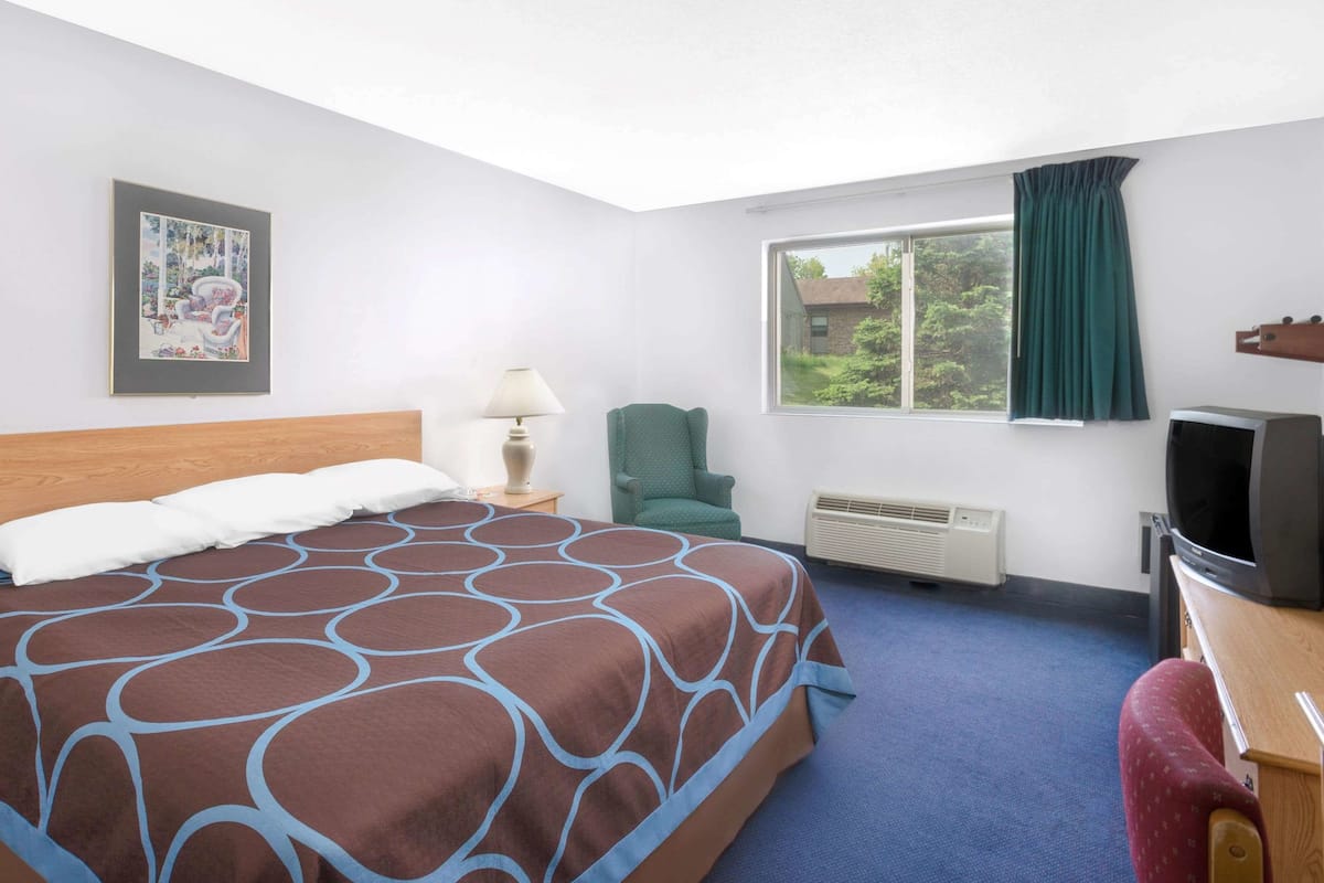 A hotel room with a bed, a chair, a TV, a window with curtains, and a picture on the wall.
