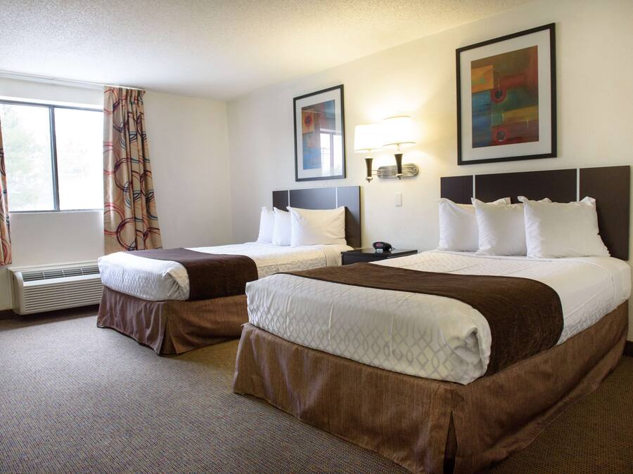 A hotel room with two beds, a large window with curtains, a wall-mounted lamp, and two framed pictures.