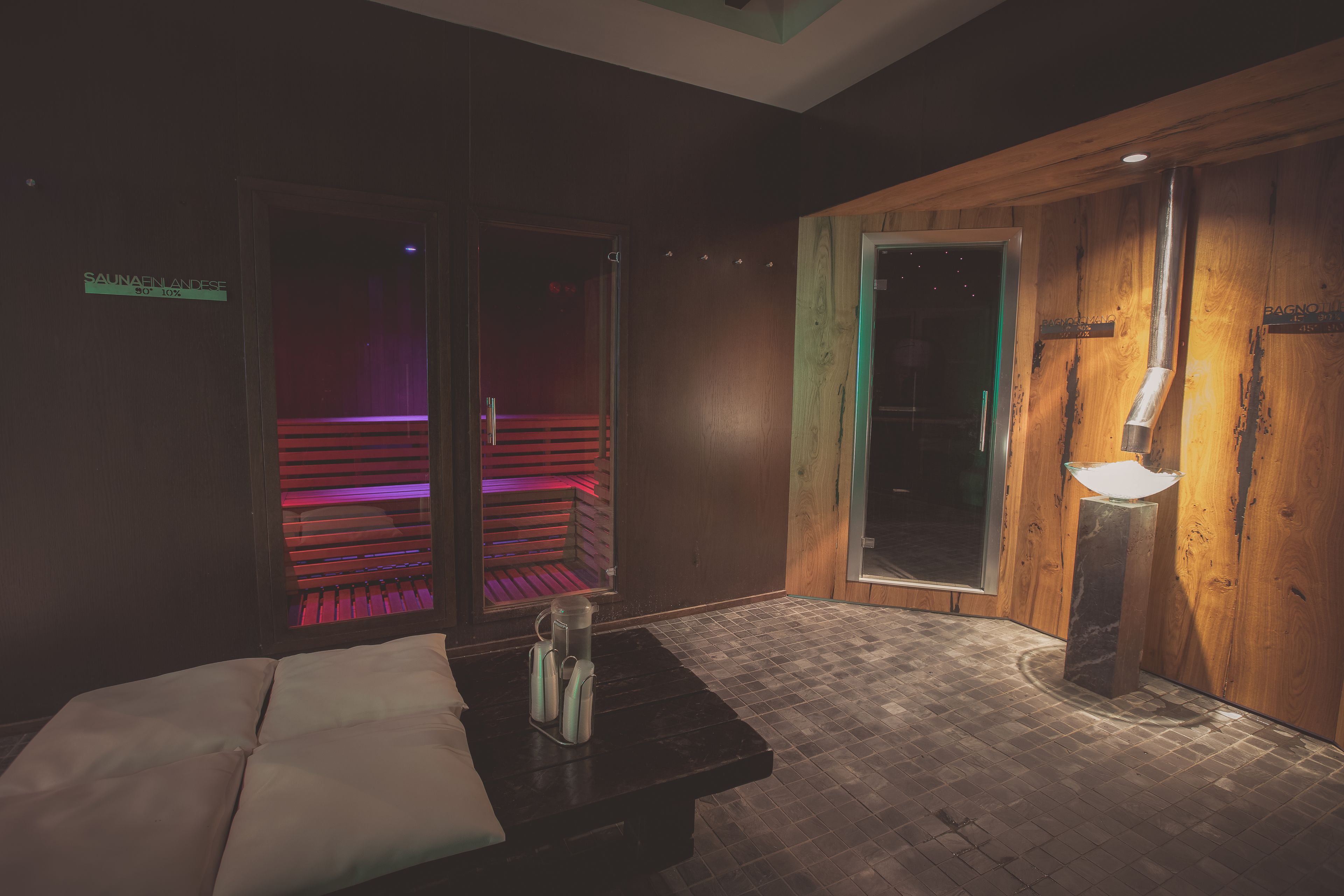 A spa room with a massage table, a wooden bench, and a glass door leading to a sauna area.