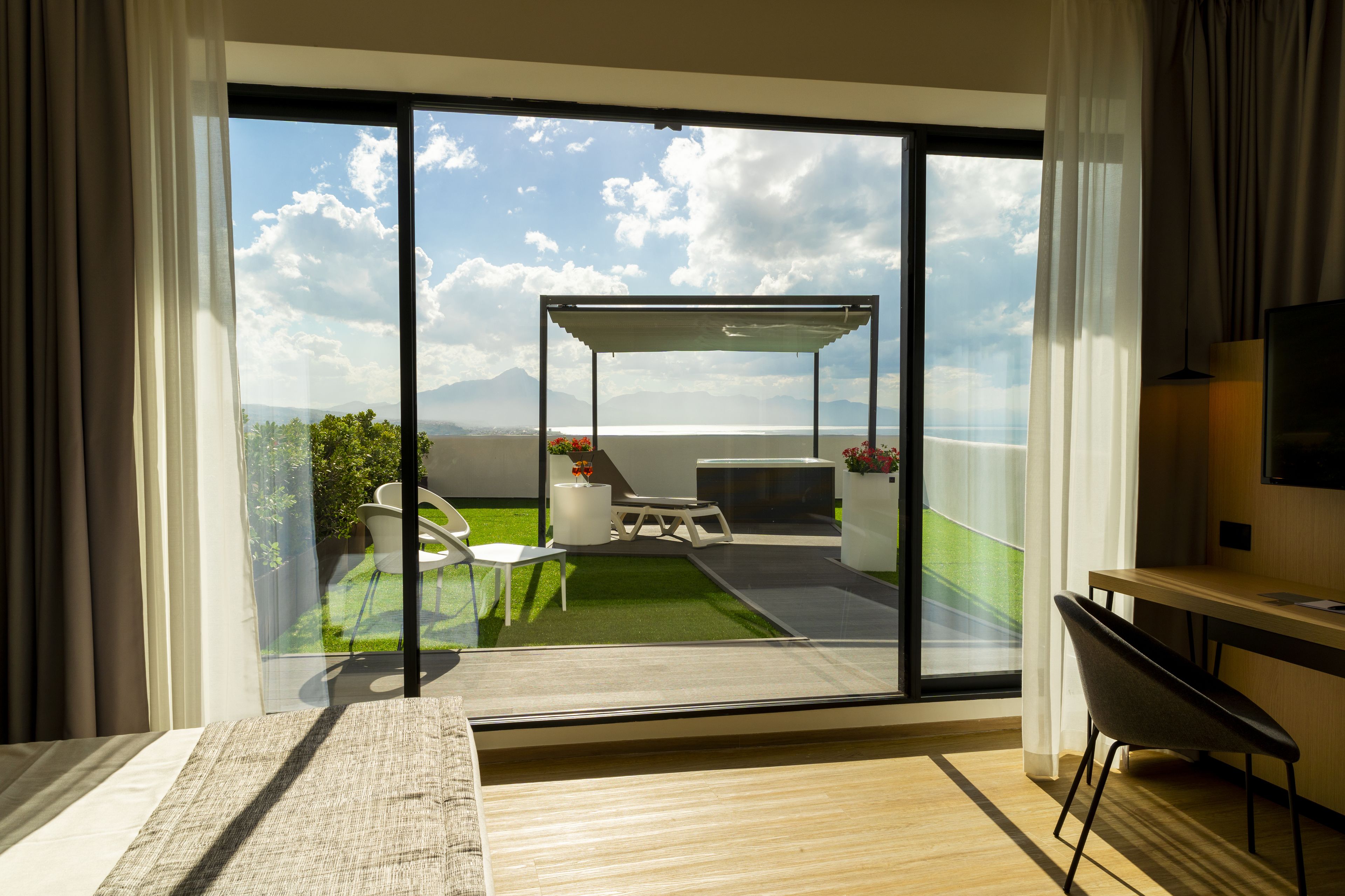 A modern hotel room with a large sliding glass door leading to a balcony with a view of the sea and mountains.