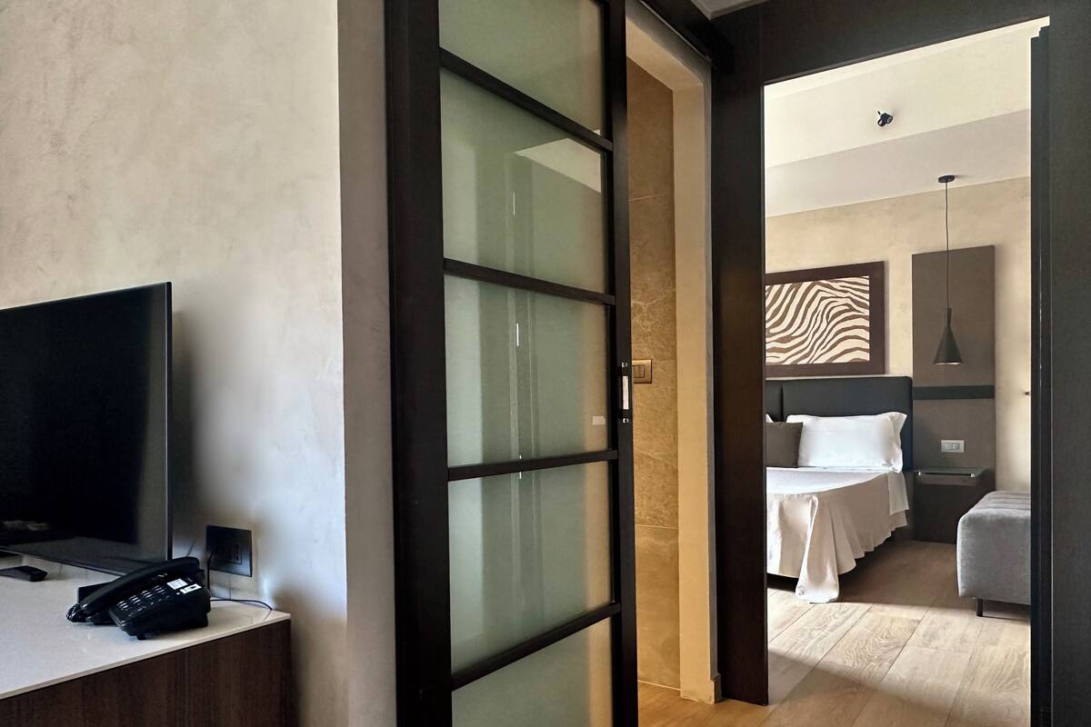 A modern hotel room with a flat-screen TV, a bedside telephone, a bed with white bedding, a zebra-patterned wall decoration, a bedside table, and a hanging pendant light.