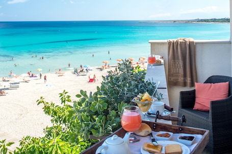 A terrace with a view of a beach, a wicker chair, a table with food and drinks, and a towel hanging on the wall.