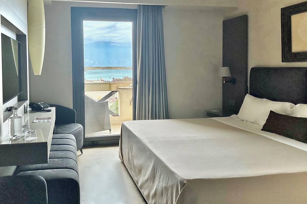 A hotel room with a bed, a desk with a phone, a chair, a TV, and a view of the sea.