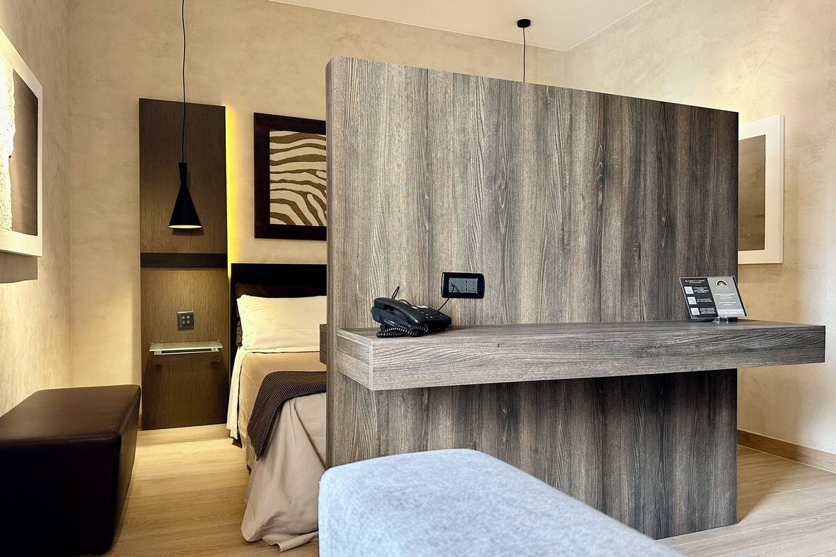 A modern hotel room with a wooden reception desk, a bed with a headboard, a bench, and a pendant light.