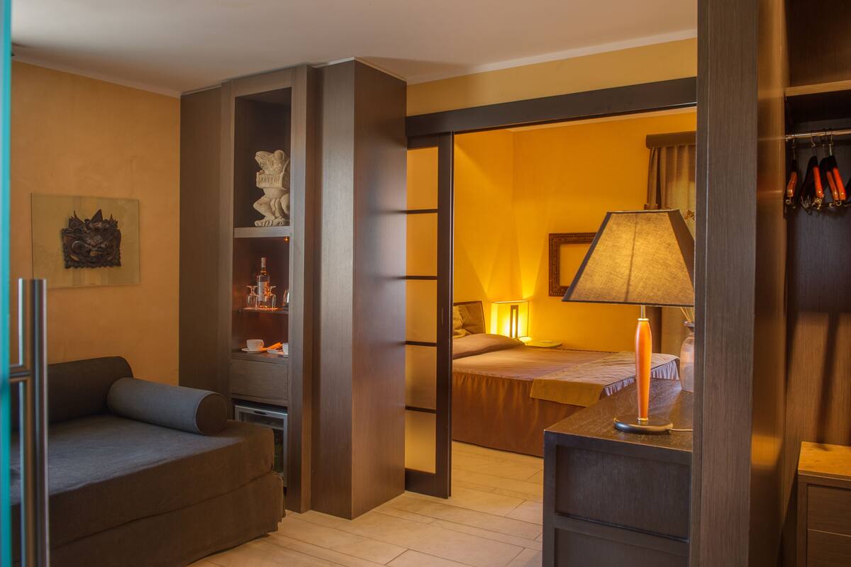 A compact hotel room with a sofa, a bed, a desk with a lamp, and a wardrobe.