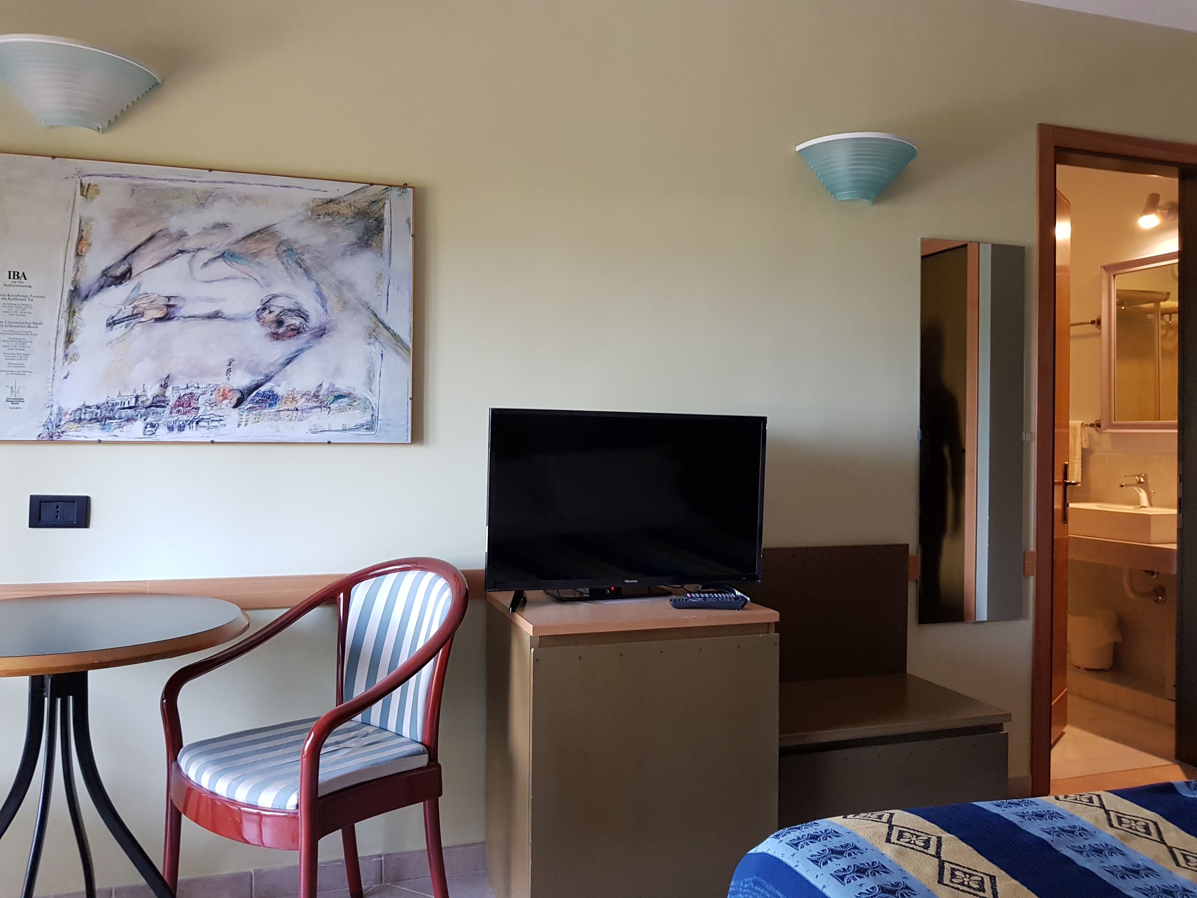 A room with a bed, a TV on a stand, a chair, a table, and a painting on the wall.