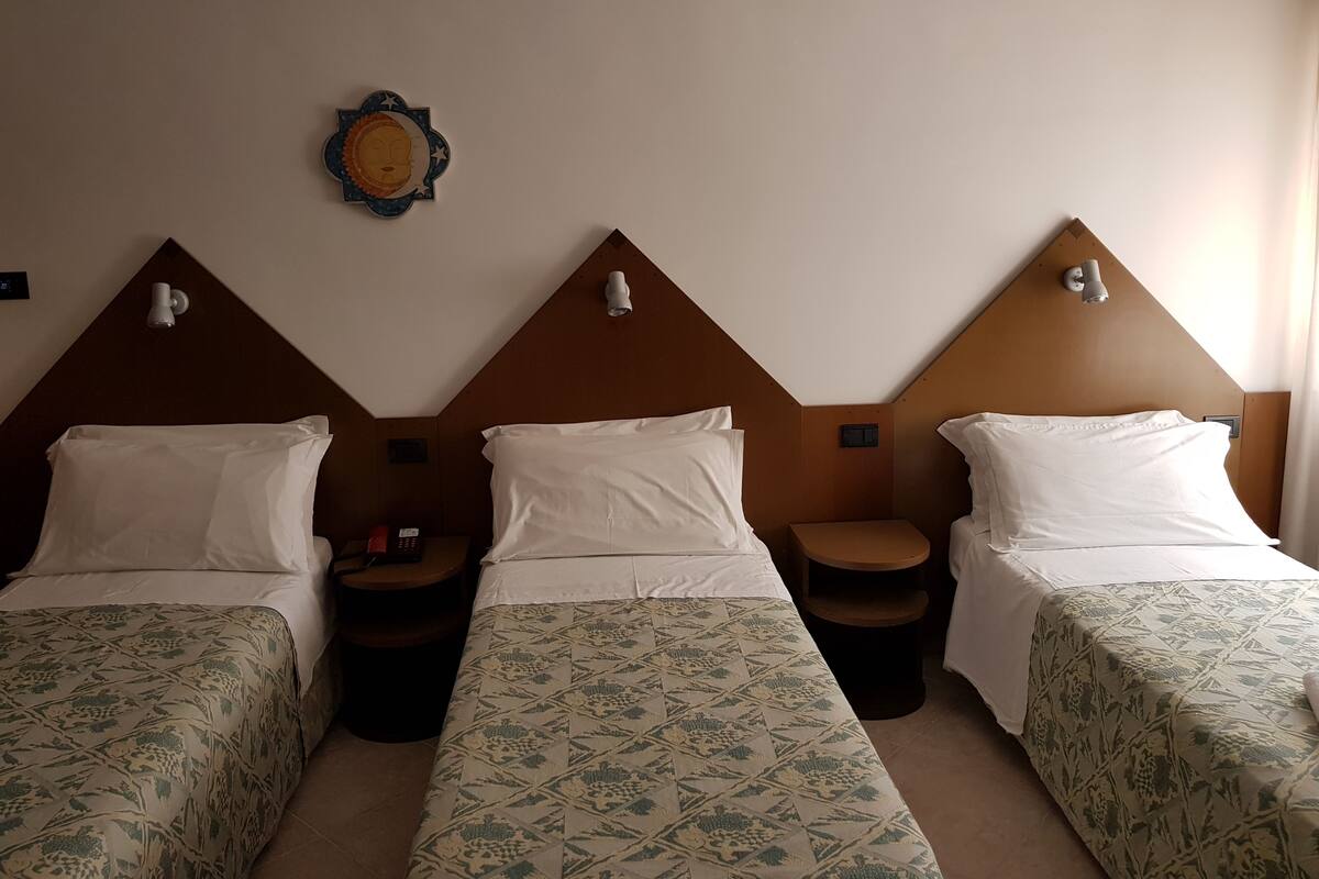 A hotel room with three single beds, each with a patterned bedspread, a small bedside table, and a wall-mounted light.