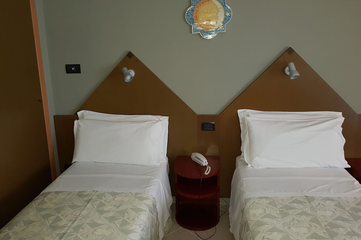 A hotel room with two beds, each with a headboard and a white bedspread. There is a small table with a telephone between the beds.