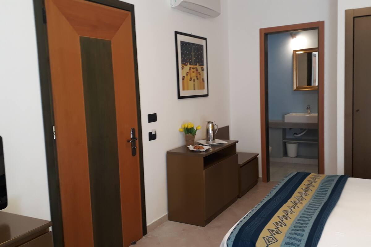 A hotel room with a bed, a desk with a kettle and flowers, a mirror, and a door.