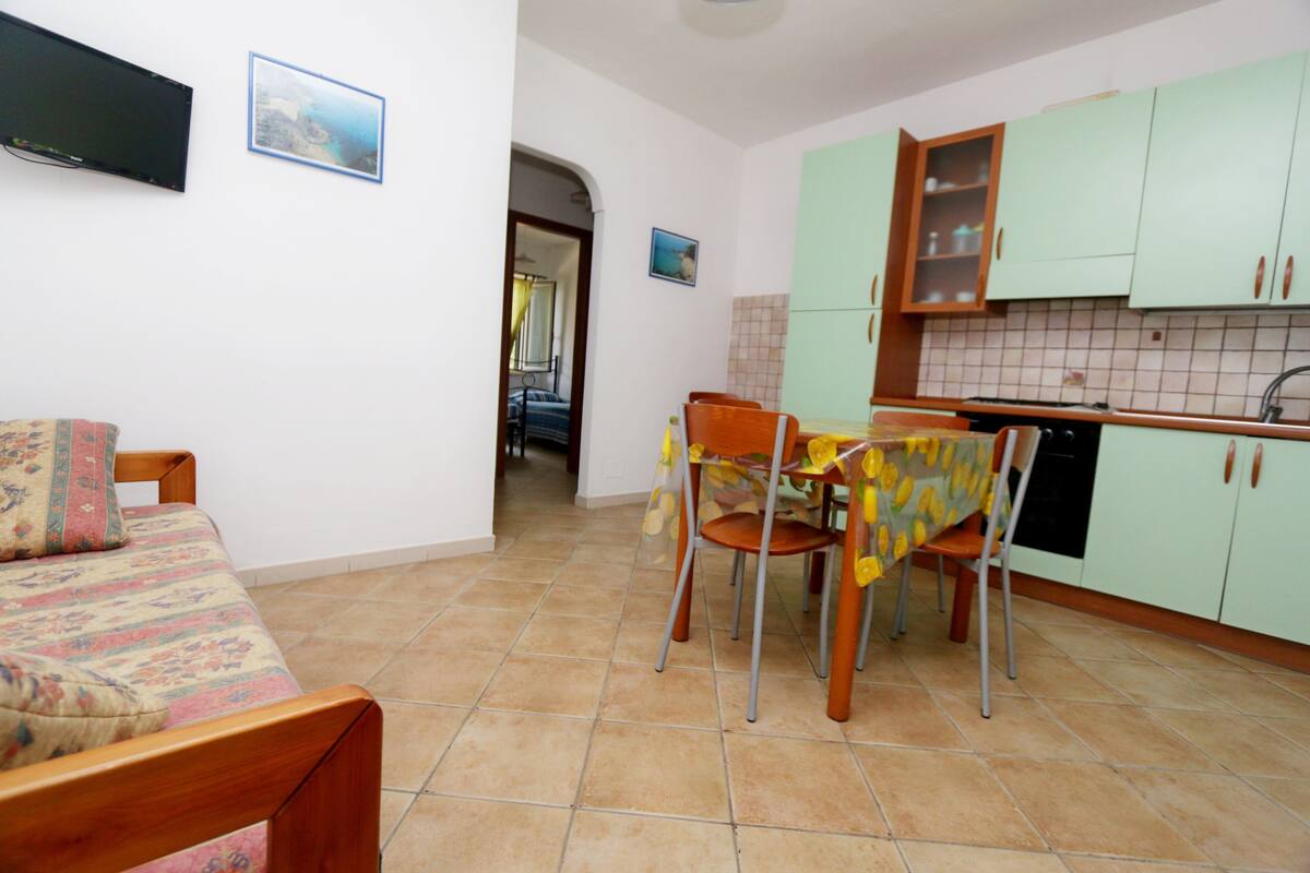 A room with a bed, a dining table with chairs, a kitchen area with cabinets, and a tiled floor.