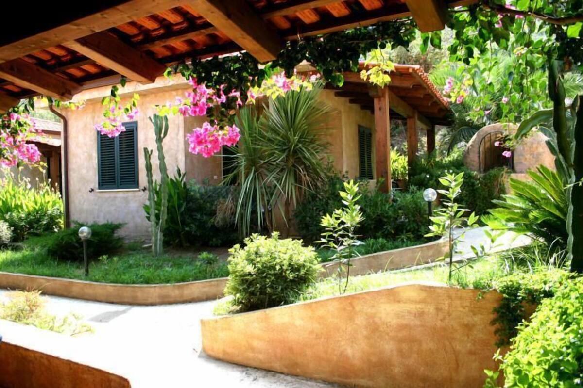 A covered patio area with a rustic wooden ceiling, surrounded by lush greenery and flowering plants.