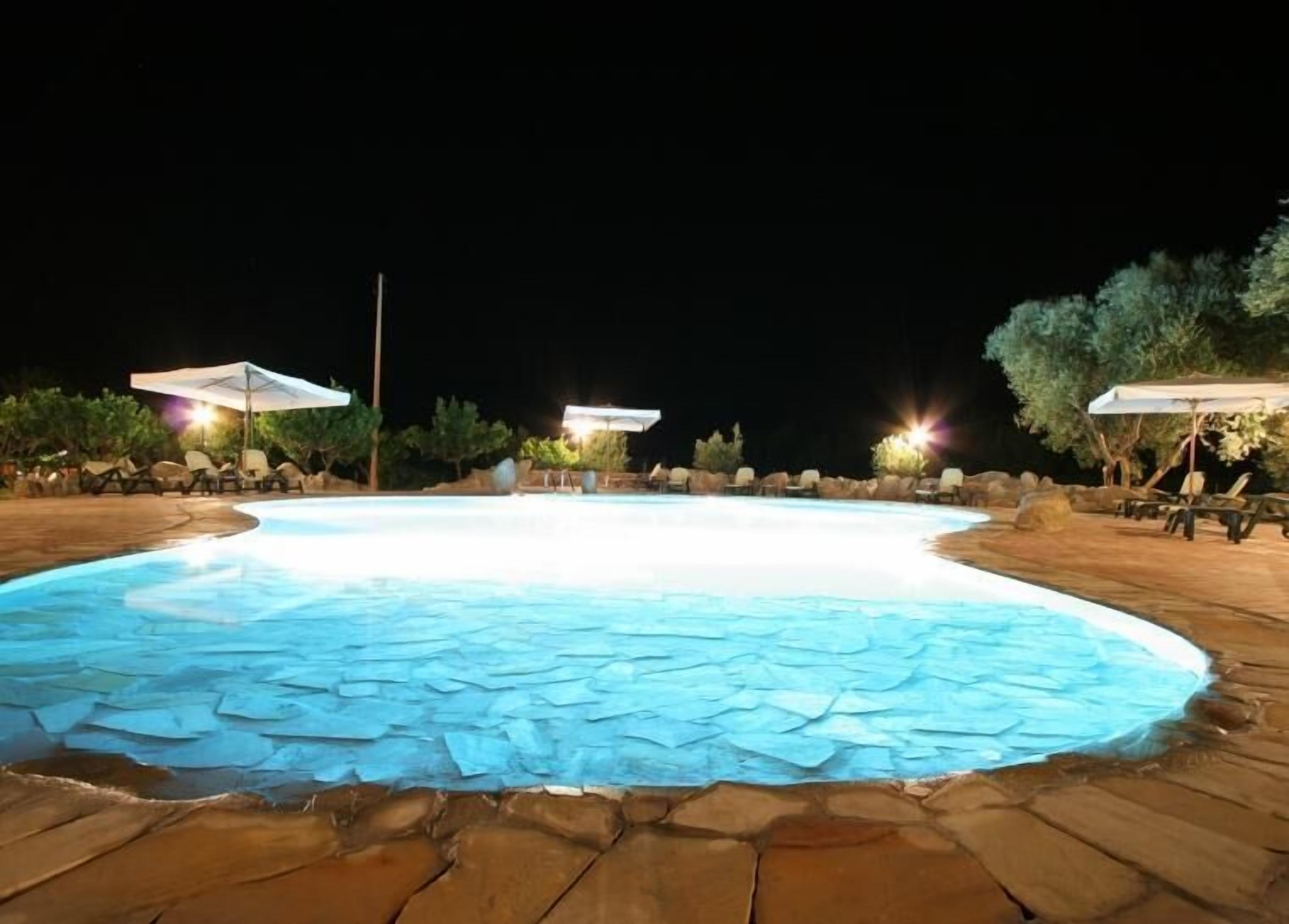 Una piscina iluminada con luz ambiental, con baldosas de piedra, rodeada de sillas de descanso y sombrillas.