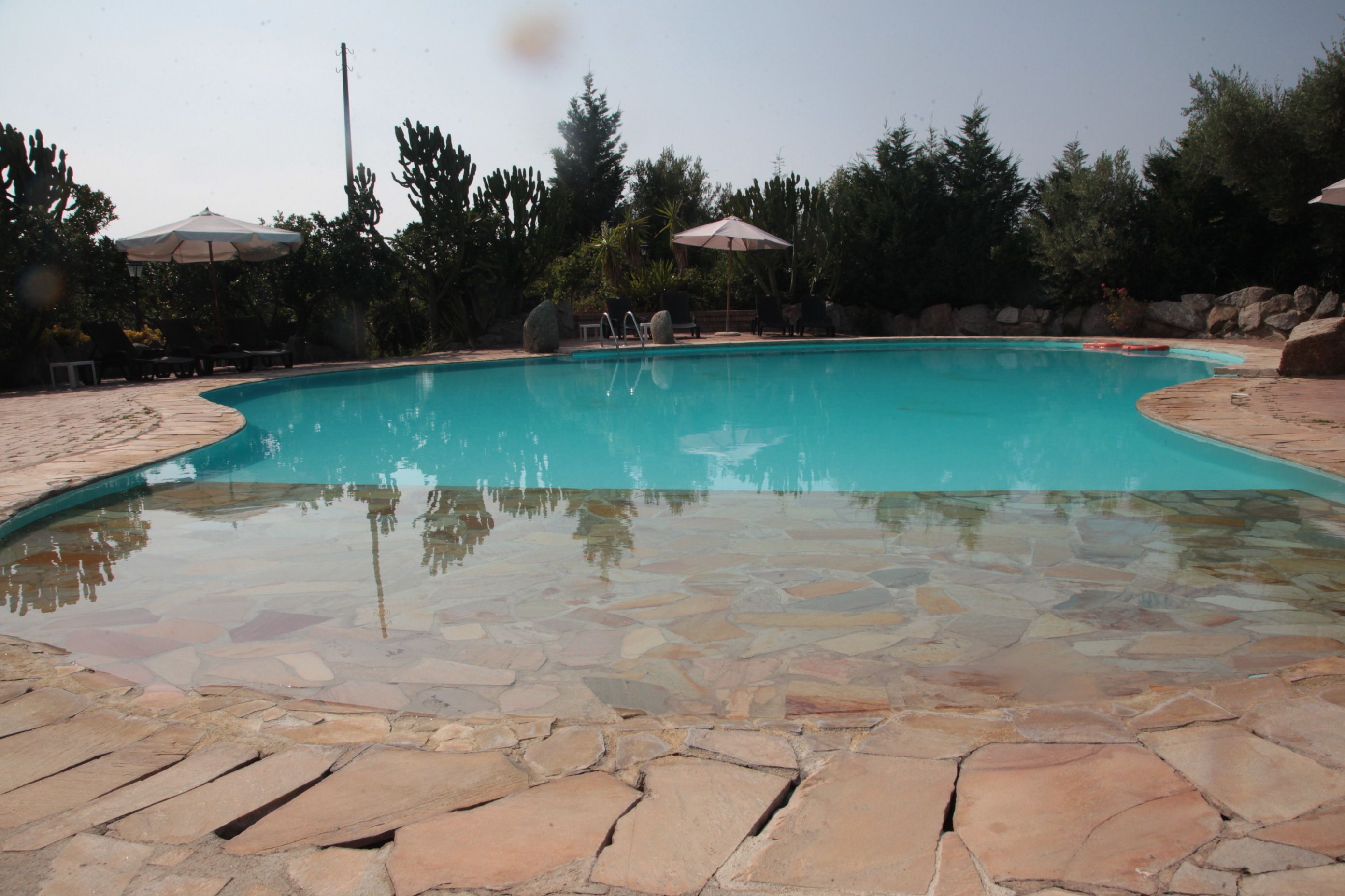 A swimming pool with stone tiles and surrounded by trees and umbrellas.