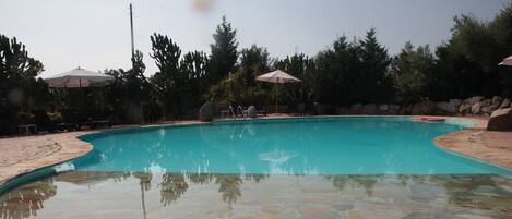 A swimming pool with stone tiles and surrounded by trees and umbrellas.