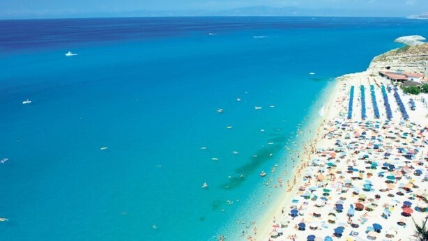 Una playa con muchos sombrillas y gente, un mar azul transparente y una costa rocosa.