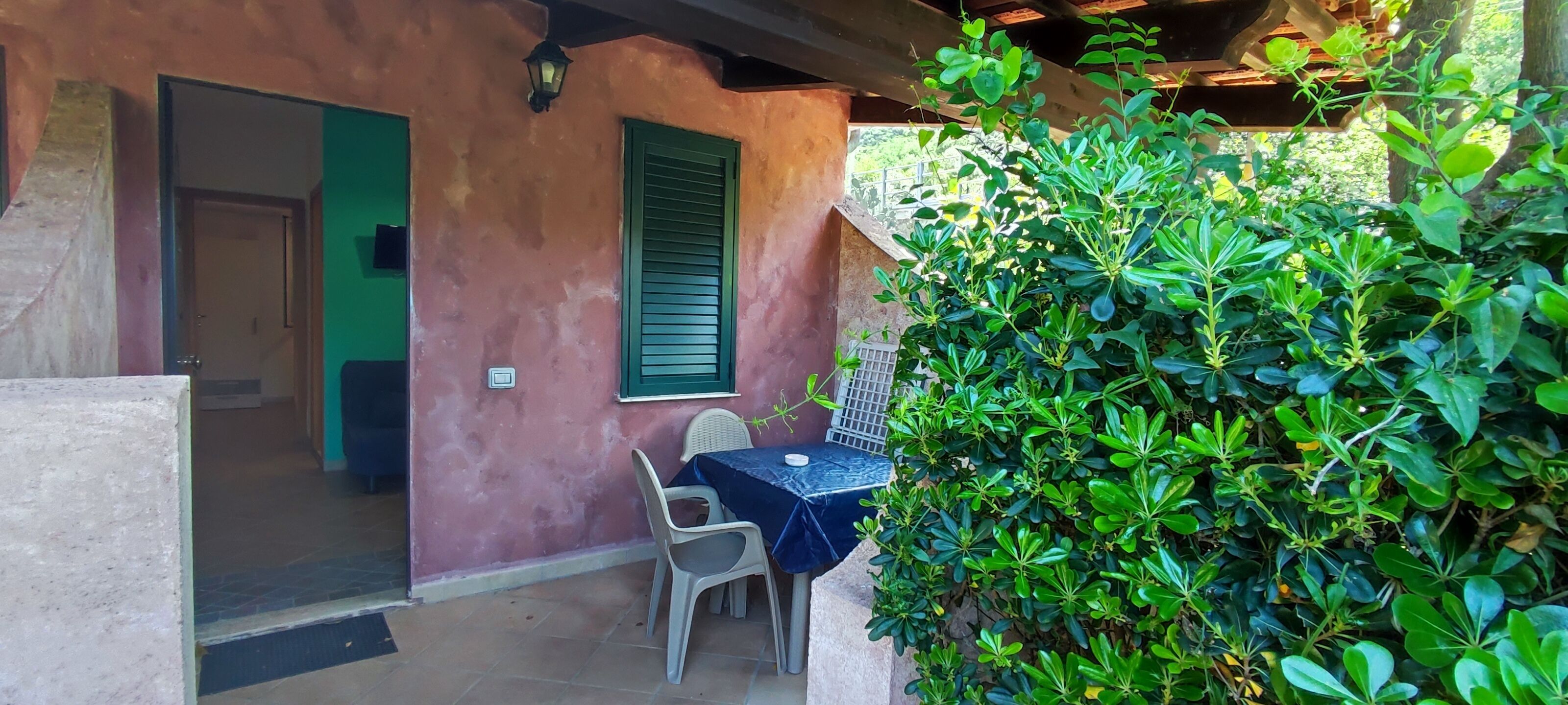 A covered outdoor seating area with a table and chairs, a door, and a wall with a green shutter.