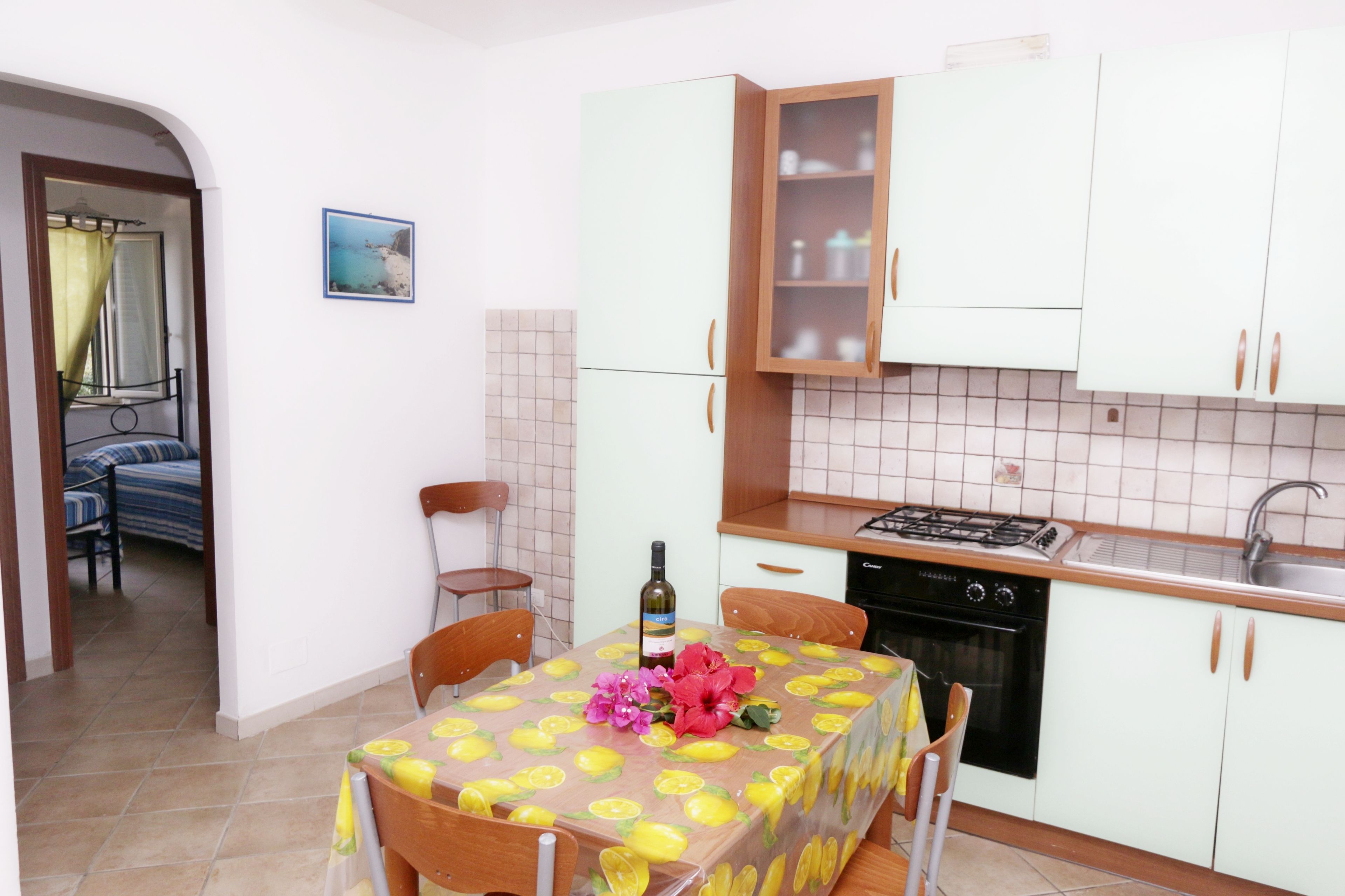 A compact kitchen with white cabinets, a gas stove, and a sink. There is a dining table with chairs and a decorative tablecloth.