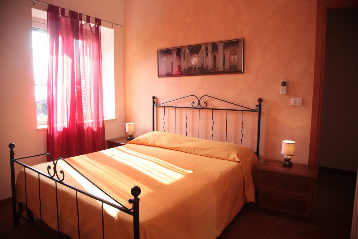 A bedroom with a bed, curtains, a nightstand, a wall-mounted lamp, and a painting.