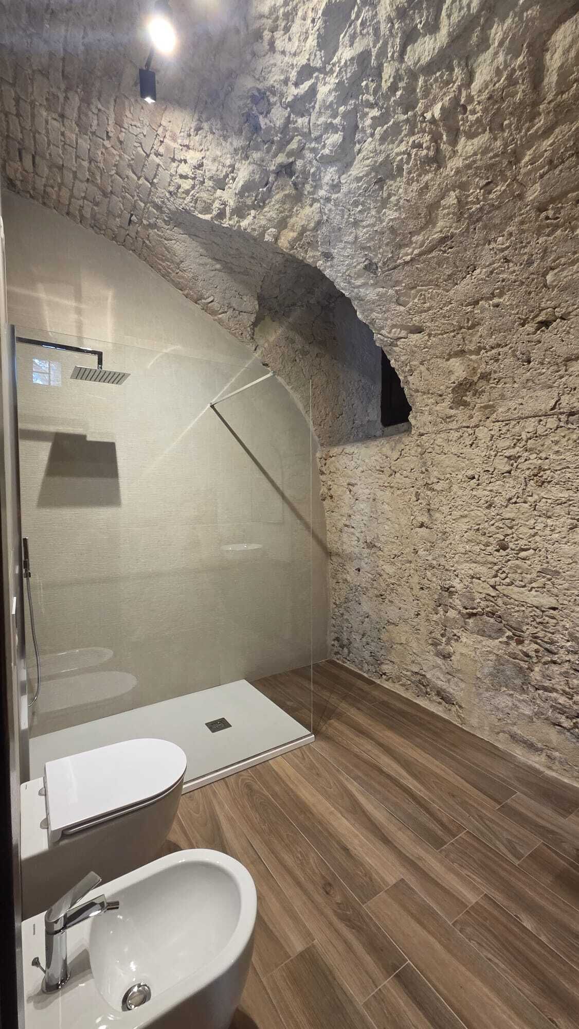 Un baño con pared de piedra, cabina de ducha de vidrio, suelo de madera y lavabo blanco.
